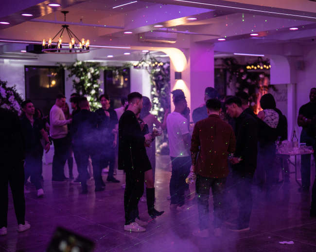 A group of people socialize in a dimly lit event space filled with purple lighting and fog effects.