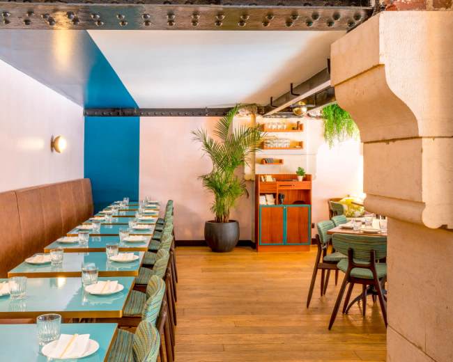 The image shows a restaurant interior with a row of tables, wooden flooring, and a corner featuring plants and shelves.