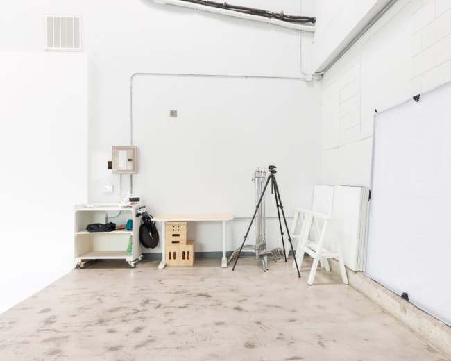 A minimalistic photo studio interior featuring a tripod, a work table, a light reflector, and bare concrete flooring.