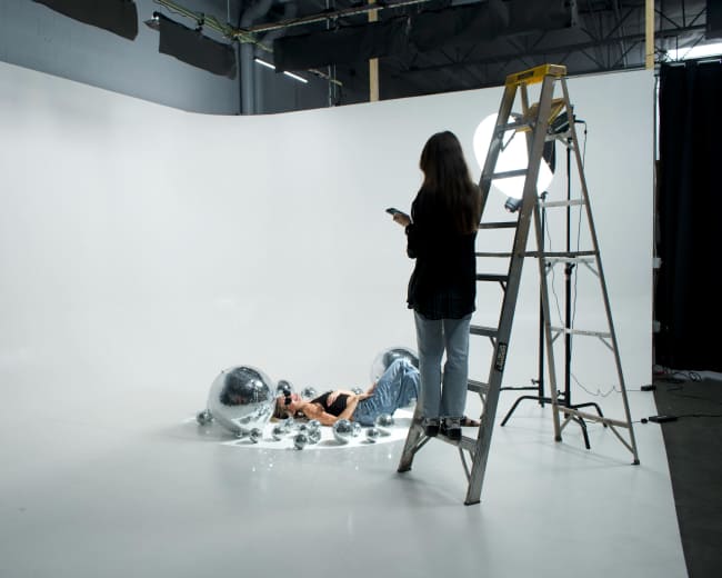 A person lies on the floor surrounded by large reflective spheres, while another person stands on a ladder holding a phone in a studio with a plain white backdrop.