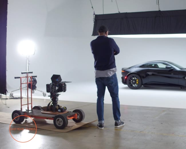 A photographer is adjusting a camera setup on a dolly while aiming at a black sports car illuminated by a large light source in a studio.