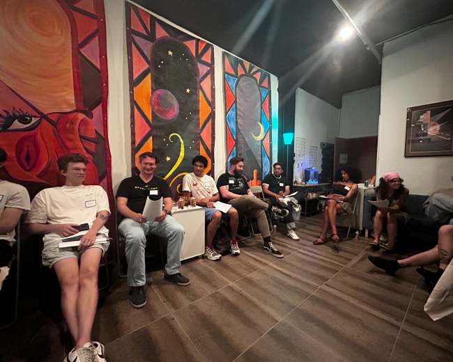 A group of people sits in a bright room adorned with colorful artwork and engaging in conversation.