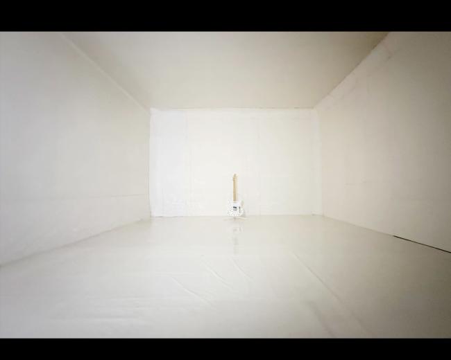 A white room with blank walls and a shiny floor contains a single electric guitar standing upright in the center.