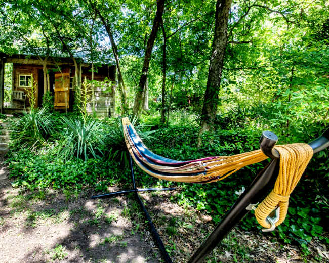 A colorful hammock is hung beside a path leading to a small cabin surrounded by lush greenery.