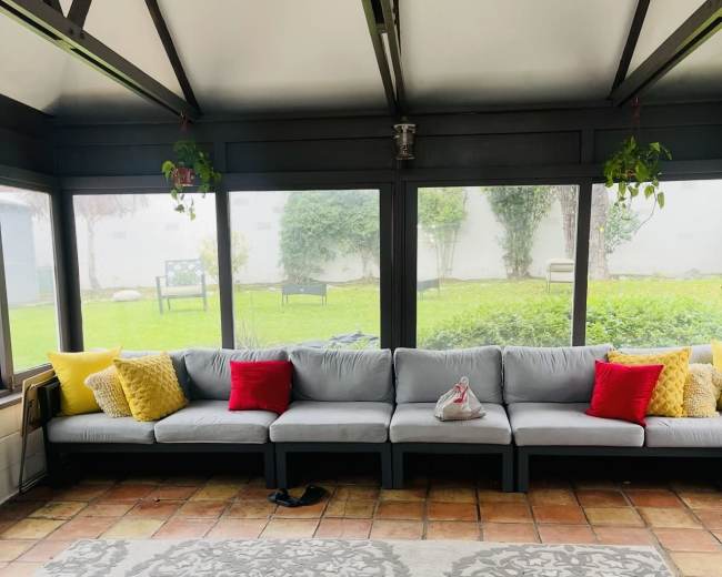 The image shows a sunroom with a long gray sectional sofa adorned with red and yellow cushions, a patterned rug, and large windows overlooking a green lawn.