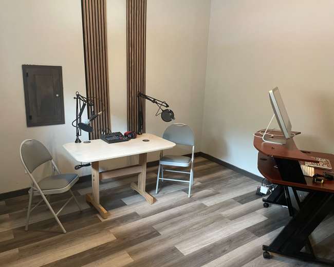 The image shows a small recording studio setup with two folding chairs, a table equipped with microphones, and a desk with a computer in a light-colored room.