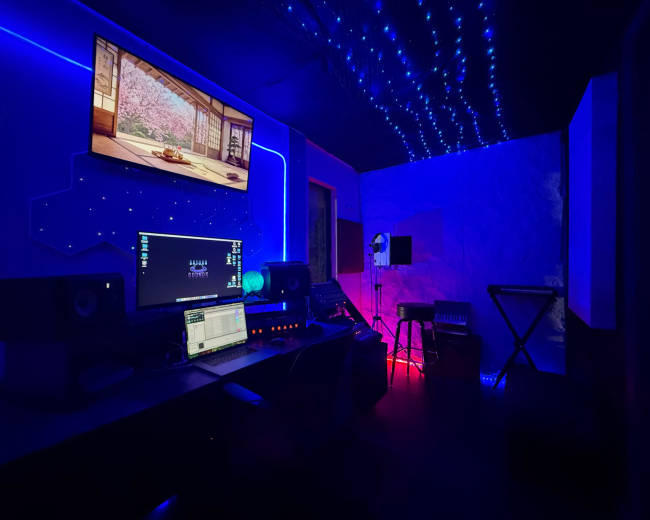The image shows a modern music studio with a computer setup, speakers, and colorful LED lights illuminating the dark space.