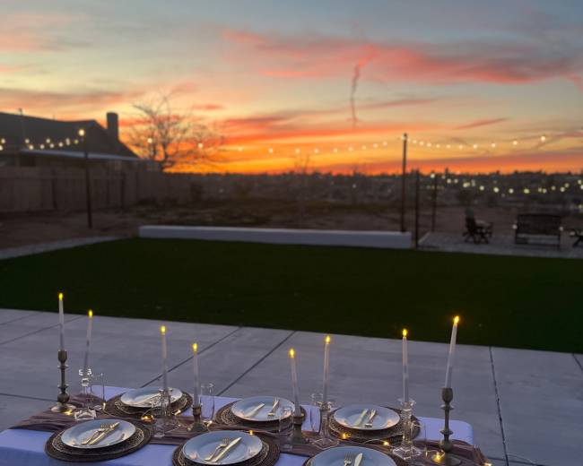 A neatly set outdoor dining table with plates, cutlery, and candles is positioned against a vibrant sunset sky.