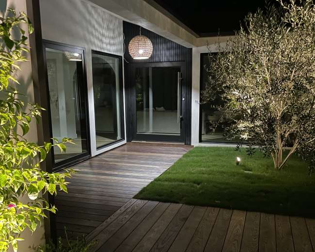 The image shows a modern outdoor area at night, featuring wooden decking, a lawn, and large glass doors illuminated by a hanging light fixture.