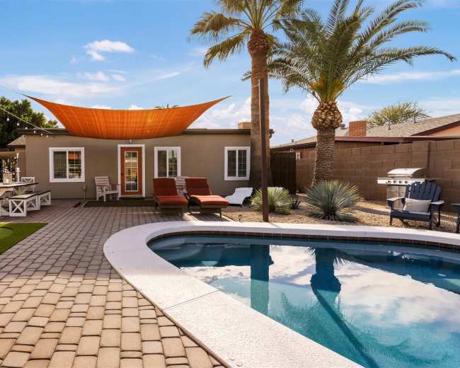 A backyard featuring a swimming pool, patio area with lounge chairs, artificial grass, and a shaded seating area, bordered by palm trees and a fence.