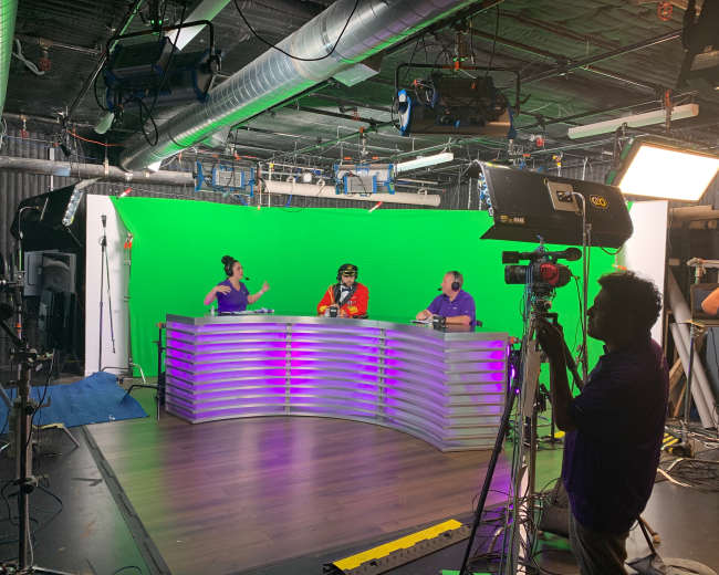 Two hosts sit at a curved desk in a studio with a green screen backdrop, while a cameraman operates a camera nearby.