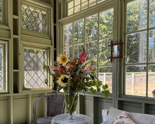 A vase of mixed flowers sits on a small table inside a sunlit room with large windows and green walls.