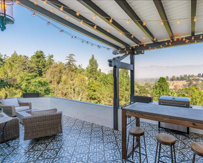 A patio area features a barbecue grill, wooden bar with stools, and string lights, overlooking a lush landscape.