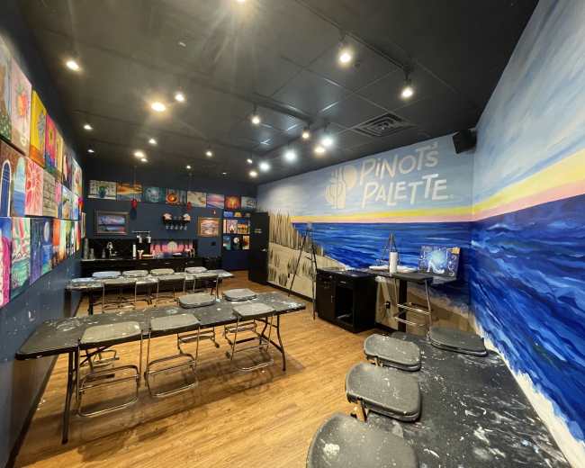 The image depicts an art studio with several tables and chairs arranged neatly, surrounded by colorful paintings on the walls.