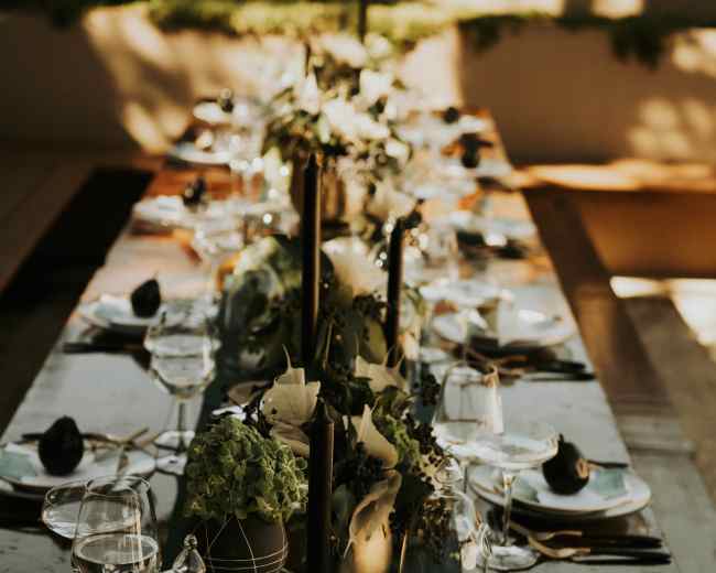 A long dining table is set with plates, glasses, and decorative centerpieces featuring greenery and candles.