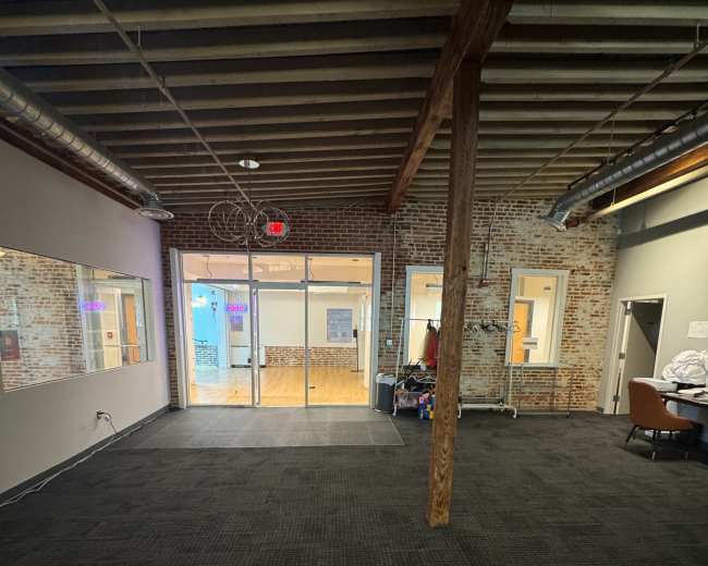 The image shows an interior view of a spacious office or studio with exposed brick walls, wooden beams, and a carpeted floor, leading to a glass entryway.