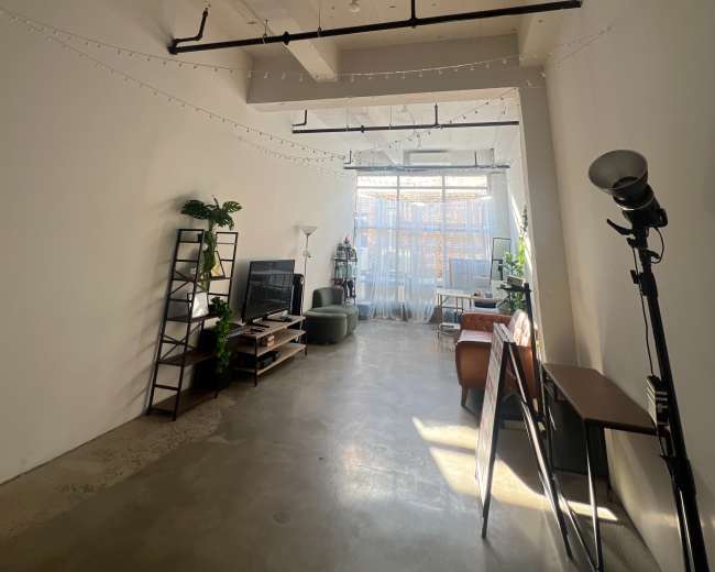 The image shows an industrial-style room with concrete floors, exposed ceiling beams, and large windows letting in natural light, filled with plants and minimalist furniture.