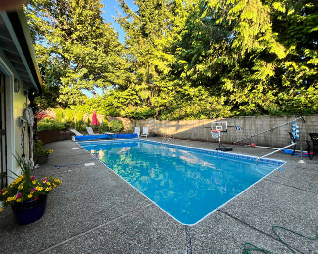 The image shows a backyard swimming pool surrounded by greenery, with lounge chairs, a basketball hoop, and a patio area.