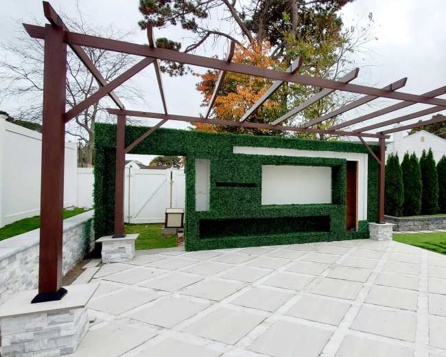The image shows a backyard patio with a pergola structure covered in greenery and a stone wall, surrounded by manicured grass and tall evergreen trees.