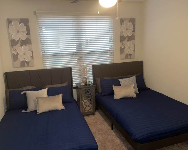 A bedroom with two twin beds covered in blue quilts, framed by light-colored walls and decorated with flower artwork above each bed.