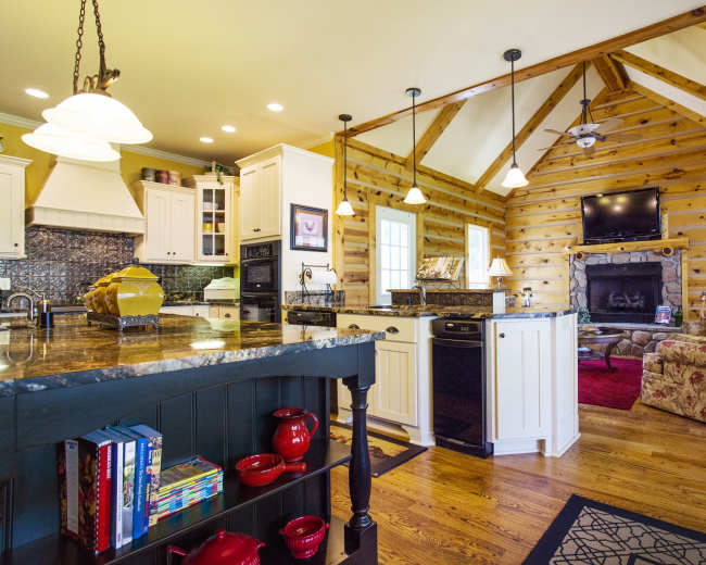 A rustic kitchen with wooden walls, an island with a dark granite countertop, and a cozy living area featuring a stone fireplace and a floral-patterned chair.
