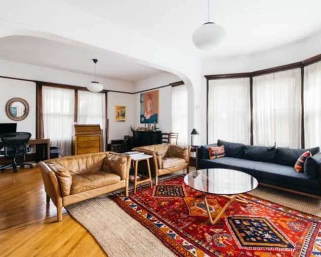 The image shows a bright living room featuring two tan leather armchairs, a dark blue sofa, a round coffee table on a colorful area rug, and large windows with sheer curtains.