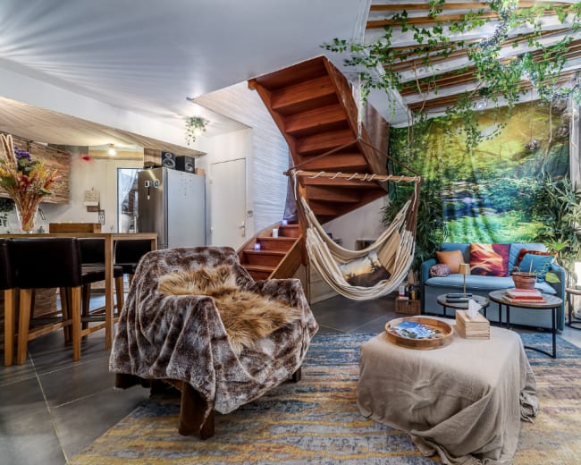 The image shows a cozy living space featuring a hammock, a staircase, and a colorful arrangement of furniture and plants.