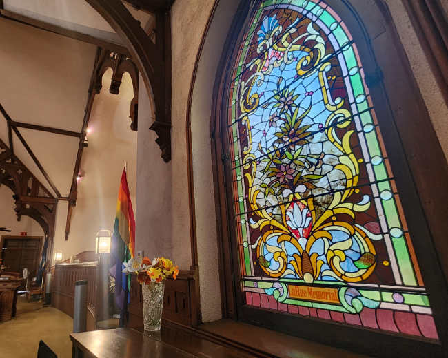 The image shows the interior of a church featuring a stained glass window, wooden beams, and flower arrangements on a table.