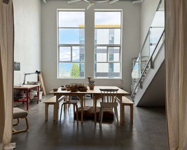 A bright, airy room features a large wooden dining table surrounded by chairs, with large windows allowing natural light and a view of an outdoor scene.