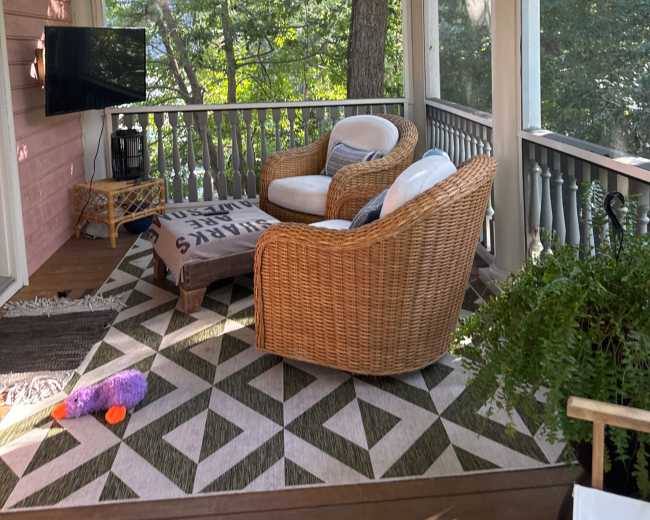 A cozy screened porch features two wicker chairs, a small table, and a television, surrounded by greenery.