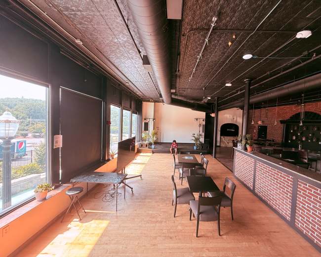 The image shows a spacious restaurant interior with tables and chairs arranged in a brightly lit area featuring large windows and industrial-style decor.