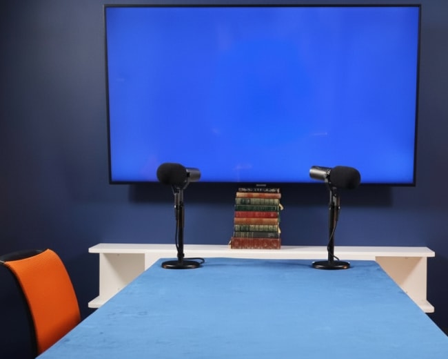 A blue wall features a large television screen, while a table with a blue cover is flanked by two chairs and topped with a stack of books and two microphones.