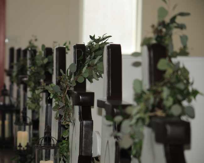 The image shows a row of wooden pews adorned with greenery and lanterns in a well-lit indoor space.