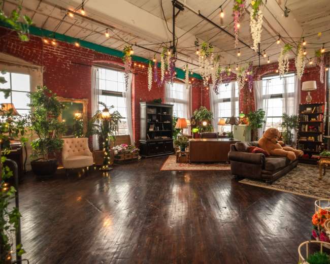The image shows a spacious, decorated room with brick walls, wooden flooring, plants, and various furniture pieces, including a sofa and armchairs.