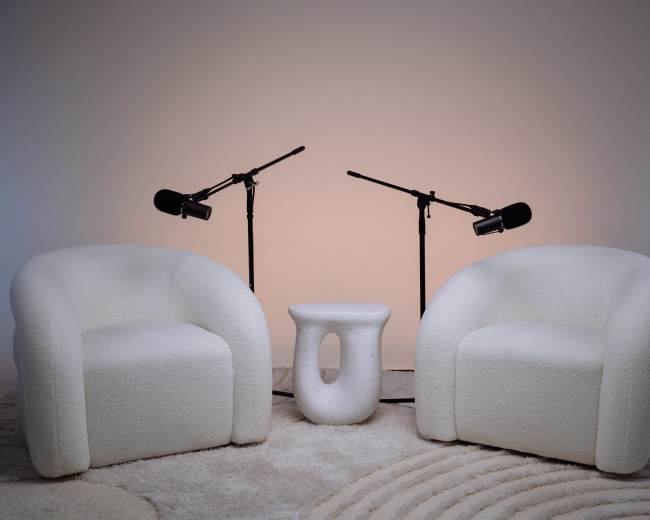Two white, plush chairs are positioned facing each other with a small, round table in between, and two microphones mounted on stands nearby.