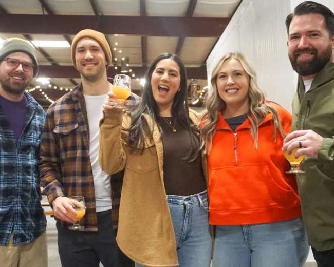 A group of five people stands together inside a venue with string lights in the background, each holding a drink and smiling at the camera.
