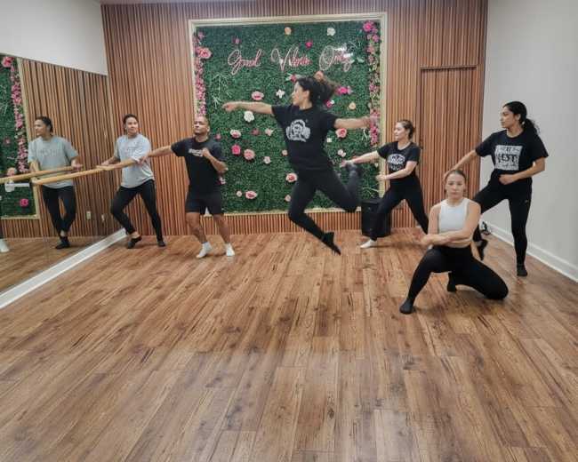 A group of dancers is practicing in a mirrored studio with a floral accent wall.
