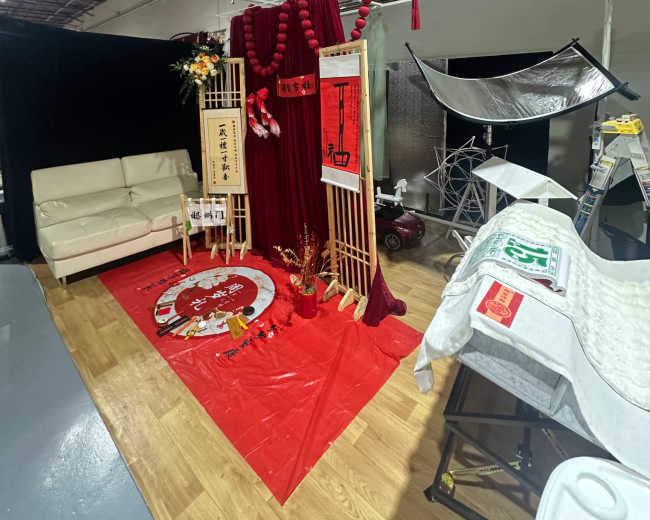 The image shows a decorated studio space featuring red fabric drapes, a white couch, and various props arranged on a wooden floor.
