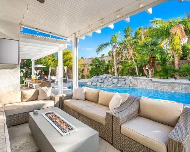 A patio area features a section of modern seating surrounding a fire table, with a swimming pool and palm trees in the background.