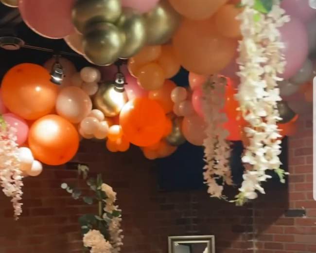 A decorated dining space with a table set for a meal, surrounded by colorful balloons and floral arrangements hanging from the ceiling.