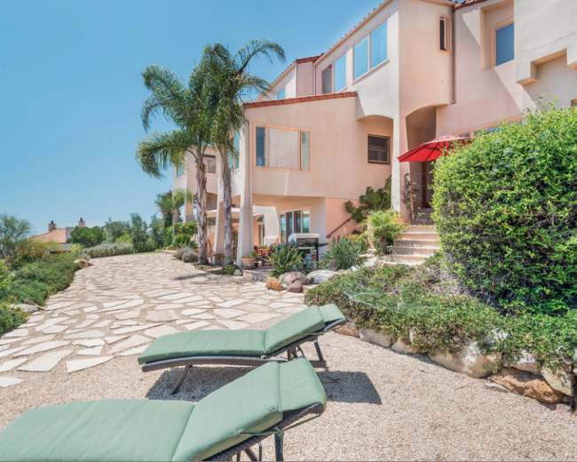 A pathway lined with palm trees and shrubs leads to a two-story stucco house with outdoor lounge chairs and an umbrella.