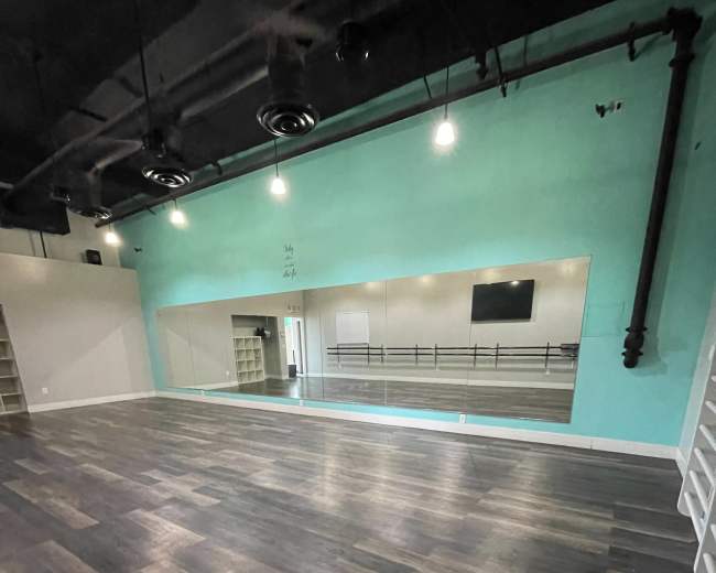 A spacious dance studio with wooden flooring, a large mirror on one wall, and teal-colored walls.