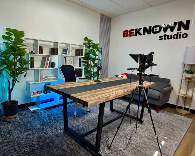 The image shows a well-organized studio space featuring a large wooden table, a microphone setup, a camera on a tripod, a comfortable gray couch, and a bookshelf filled with books and plants against the wall.
