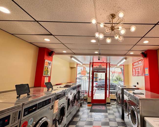 The interior of a laundromat features rows of washer and dryer machines, a seating area, and large windows displaying a view of the outside.