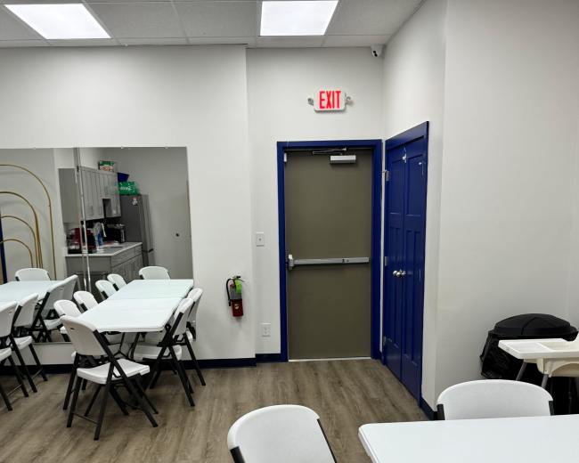 The image shows a room with several white folding tables and chairs arranged for seating, featuring a kitchenette area and an exit door at the rear.