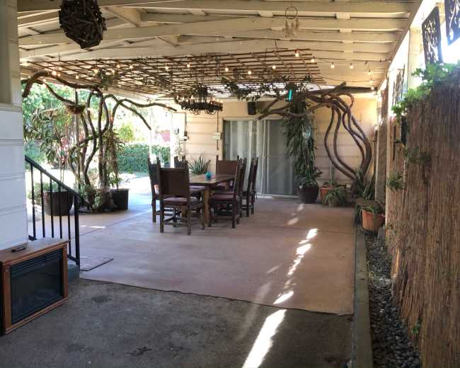 The image shows a covered outdoor patio area with a wooden table and chairs surrounded by greenery and decorative elements.