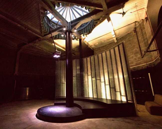 The image shows a dimly lit circular room with a central circular stage and tall, translucent panels reflecting light.