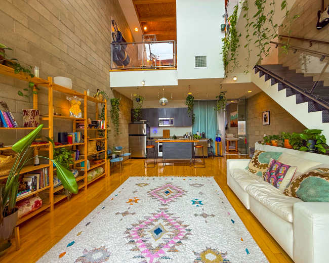 The image shows a modern living room with a white sofa, a large patterned rug, and a staircase leading to an upper level, surrounded by plants and wooden shelving.