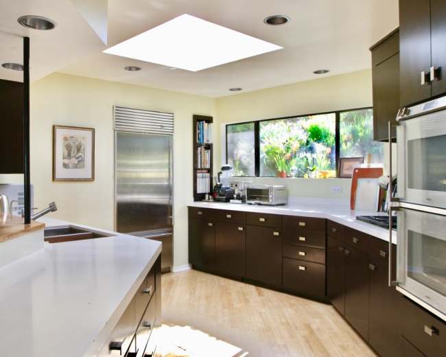 The image depicts a modern kitchen with sleek cabinetry, a skylight overhead, and large windows offering a view of greenery outside.