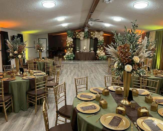 The image shows a decorated indoor event space with elegantly set tables featuring golden chairs, floral centerpieces, and a backdrop of balloons.